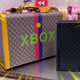 The Gucci Xbox has been placed in front of the Gucci monogram trunk on a table.